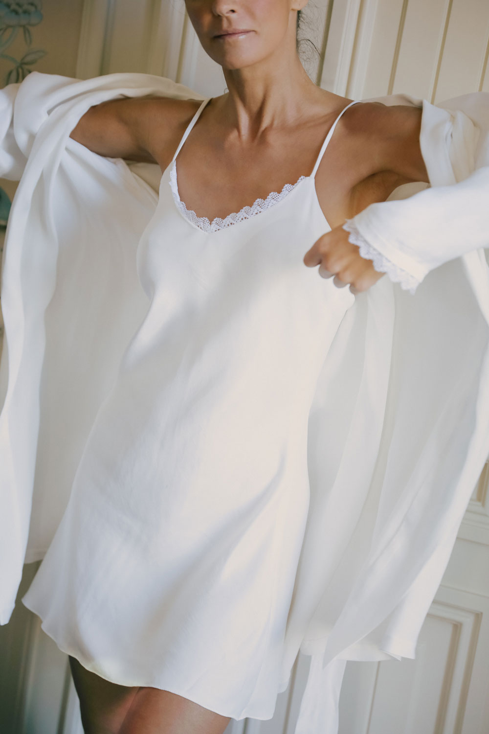 woman standing, dressed in a white silk nightgown from Leizi's collection, putting on the robe from the same collection