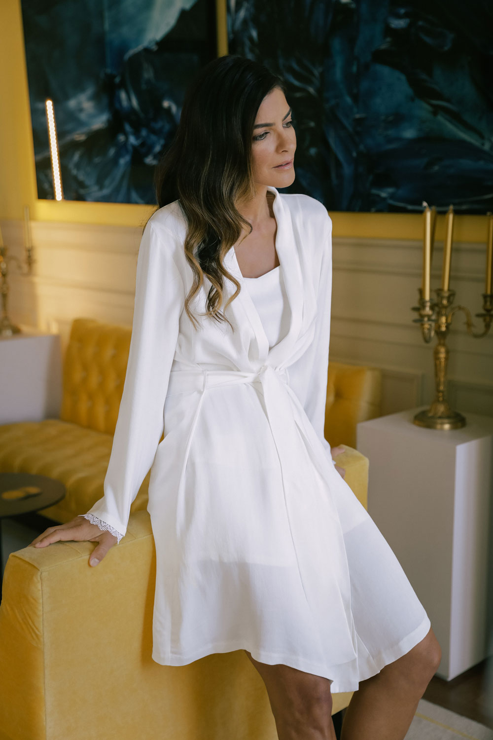 woman leaning against a yellow sofa, dressed in a white silk robe from Leizi's Silky Dreams collection.