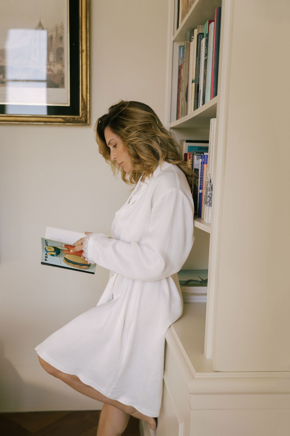 woman seen from the side, leaning against a bookcase, reading, dressed in a white silk robe from Leizi's Silky Dreams collection.
